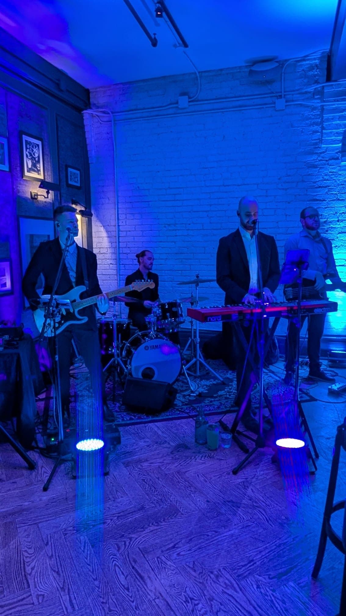 Supersonic Hearts playing in a room washed in blue light, spotlights on the floor in front of them and the gallery wall behind.