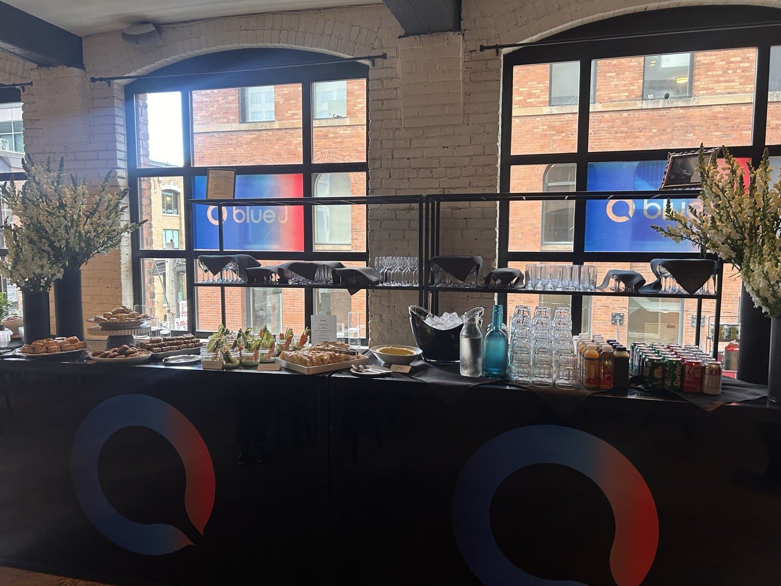 The bar set for a break, its front wrapped in two large Blue J marks, with pastries, crudité jars, juice and water on top and Blue J graphics in the arched windows behind.