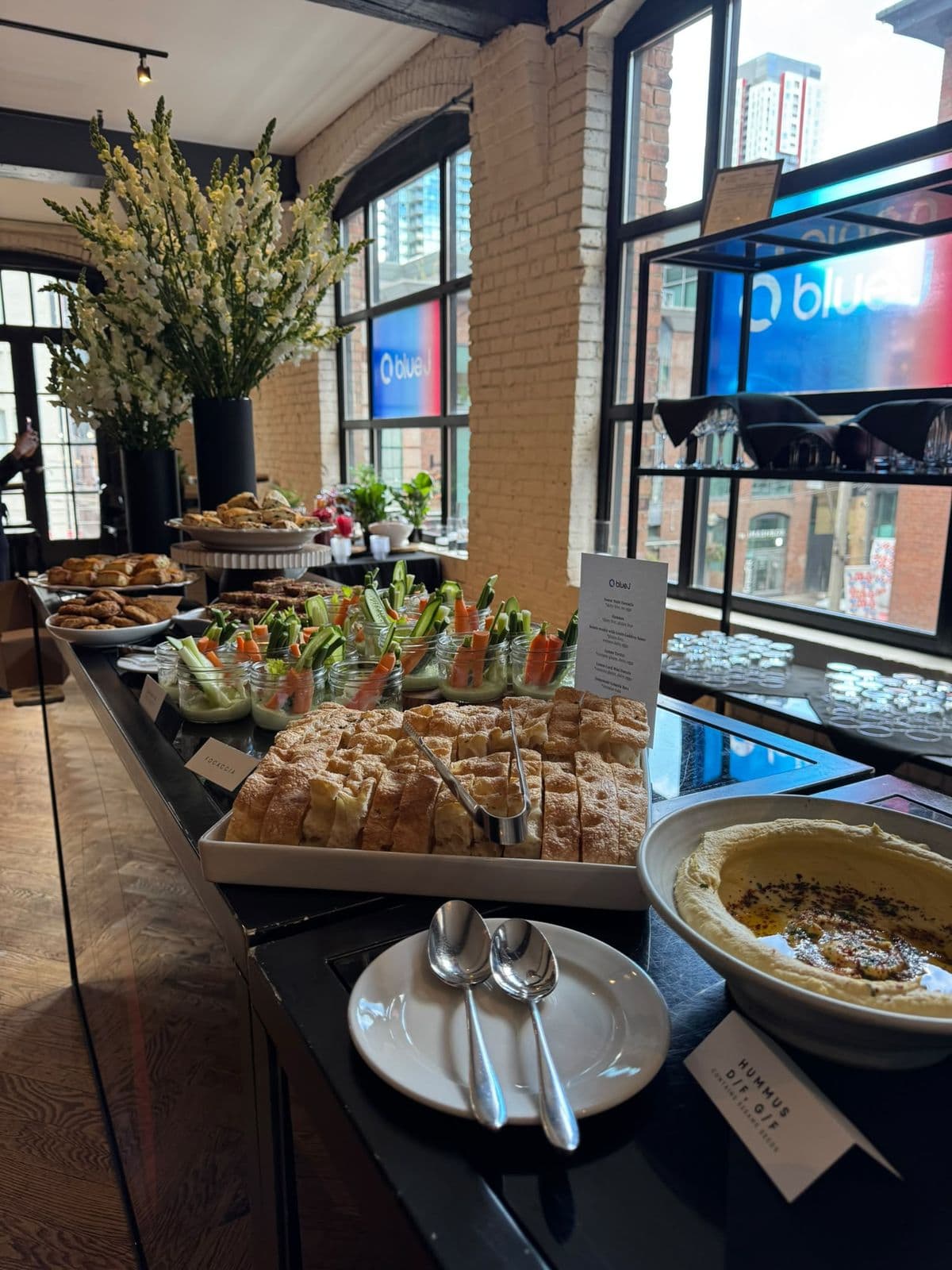 The afternoon break laid out along the bar: focaccia, hummus, crudité jars and pastries, with white flowers and Blue J graphics in the windows.