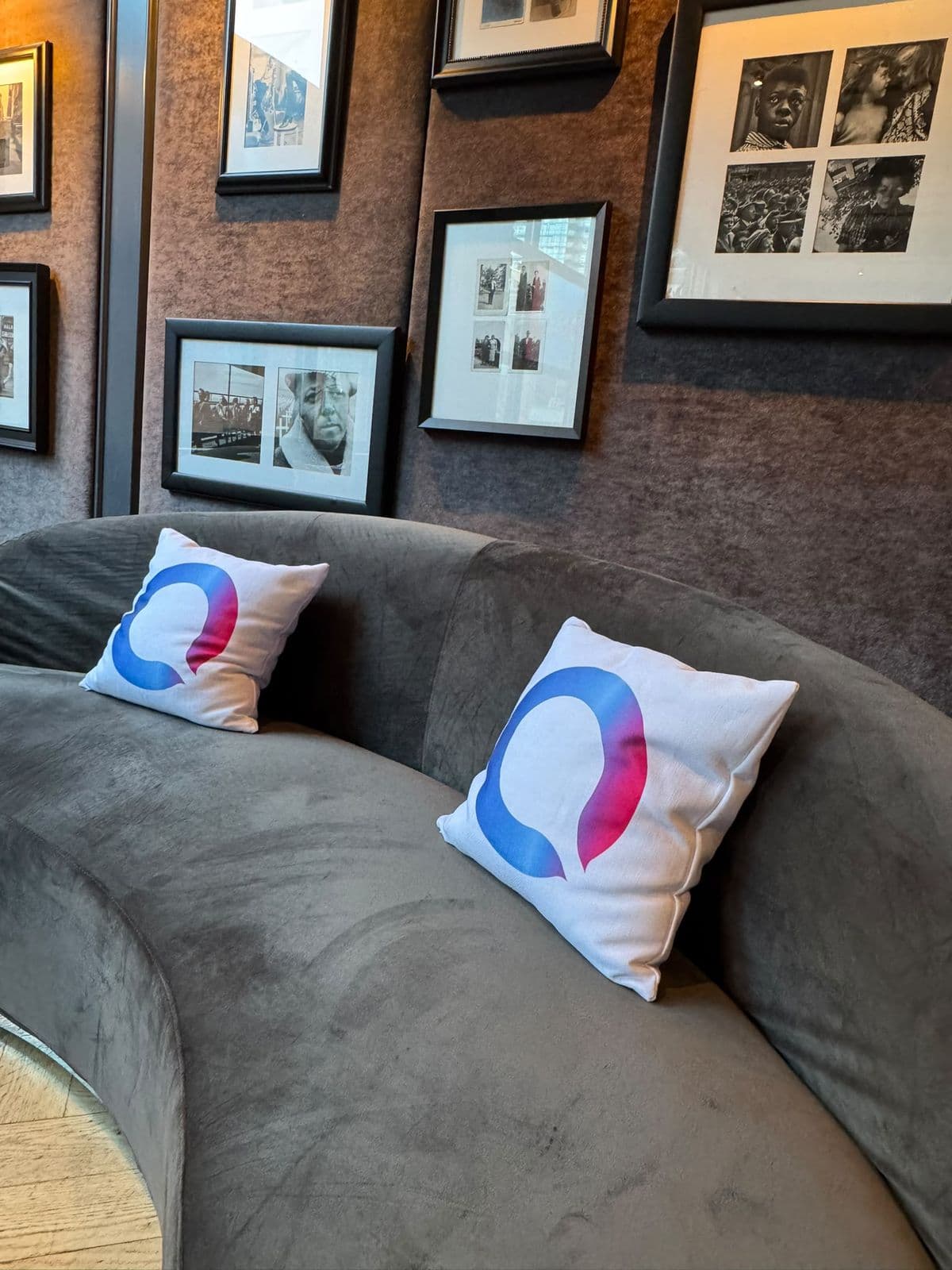 Two white cushions with the Blue J mark on a grey velvet sofa, framed black and white photographs on the wall behind.