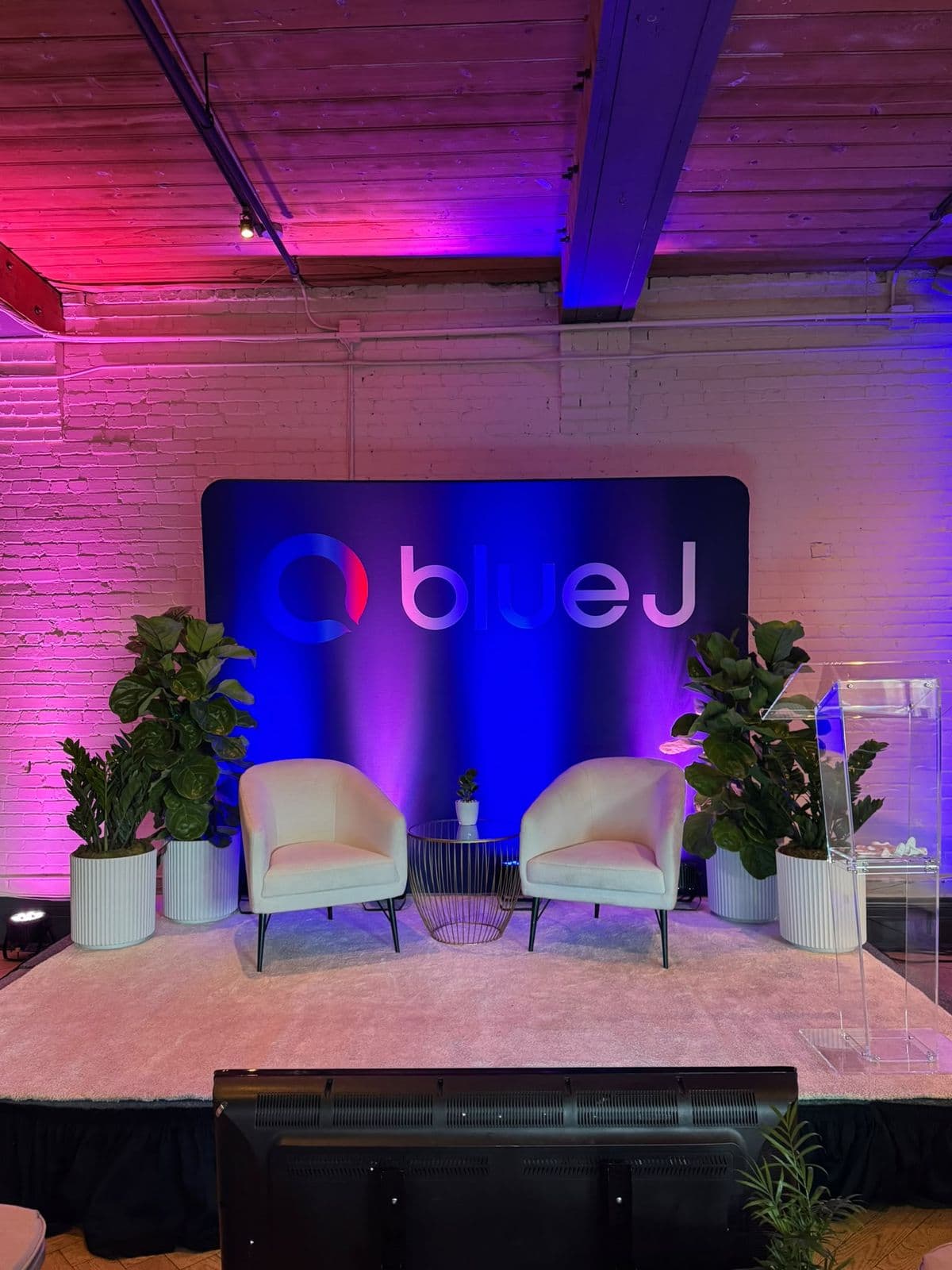 A low stage set for the sessions: a Blue J backdrop lit in pink and blue, two cream armchairs and a clear lectern, with plants on either side, against white brick.
