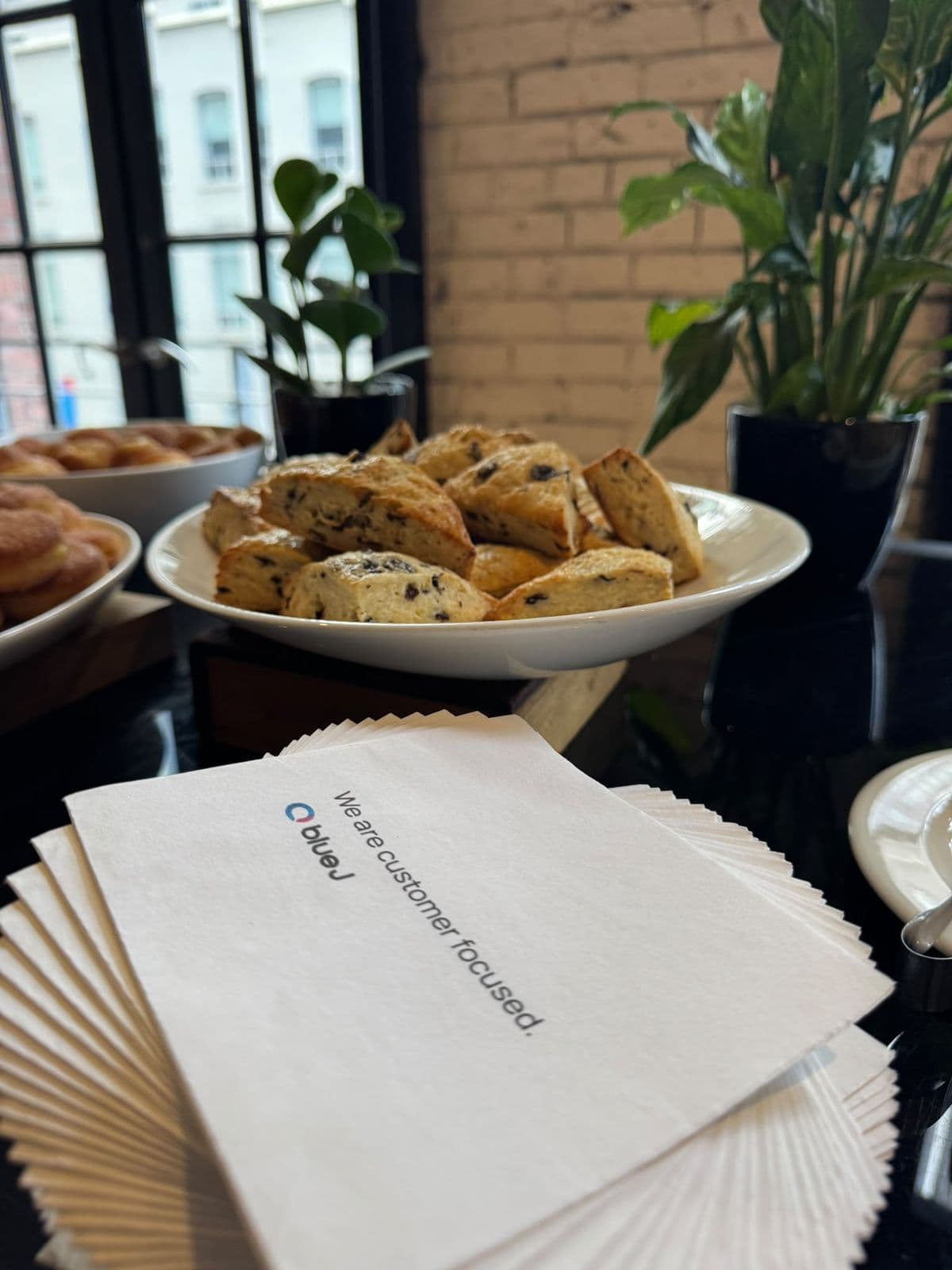 A stack of white napkins printed with the Blue J logo and “We are customer focused.”, beside a bowl of scones.