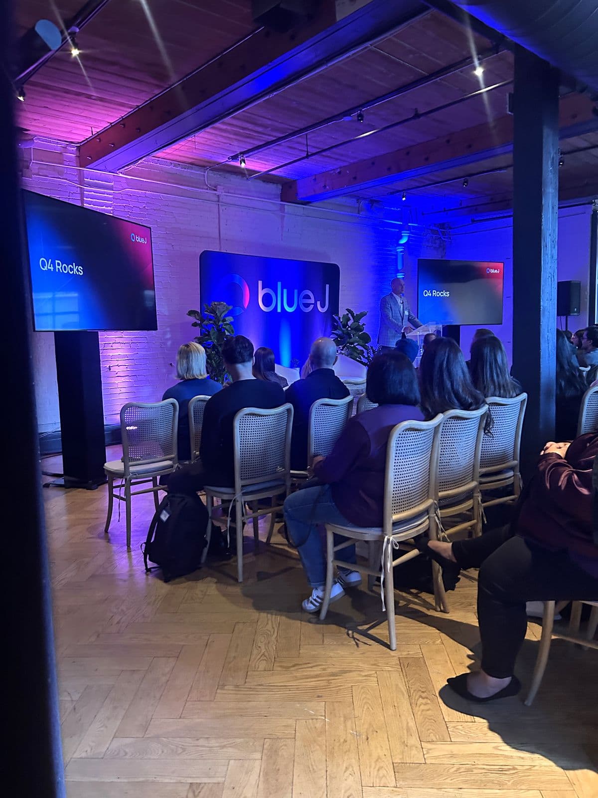 A session in progress: a speaker at the lectern in front of the Blue J backdrop, screens on either side, and the audience seated in rows, seen from behind.
