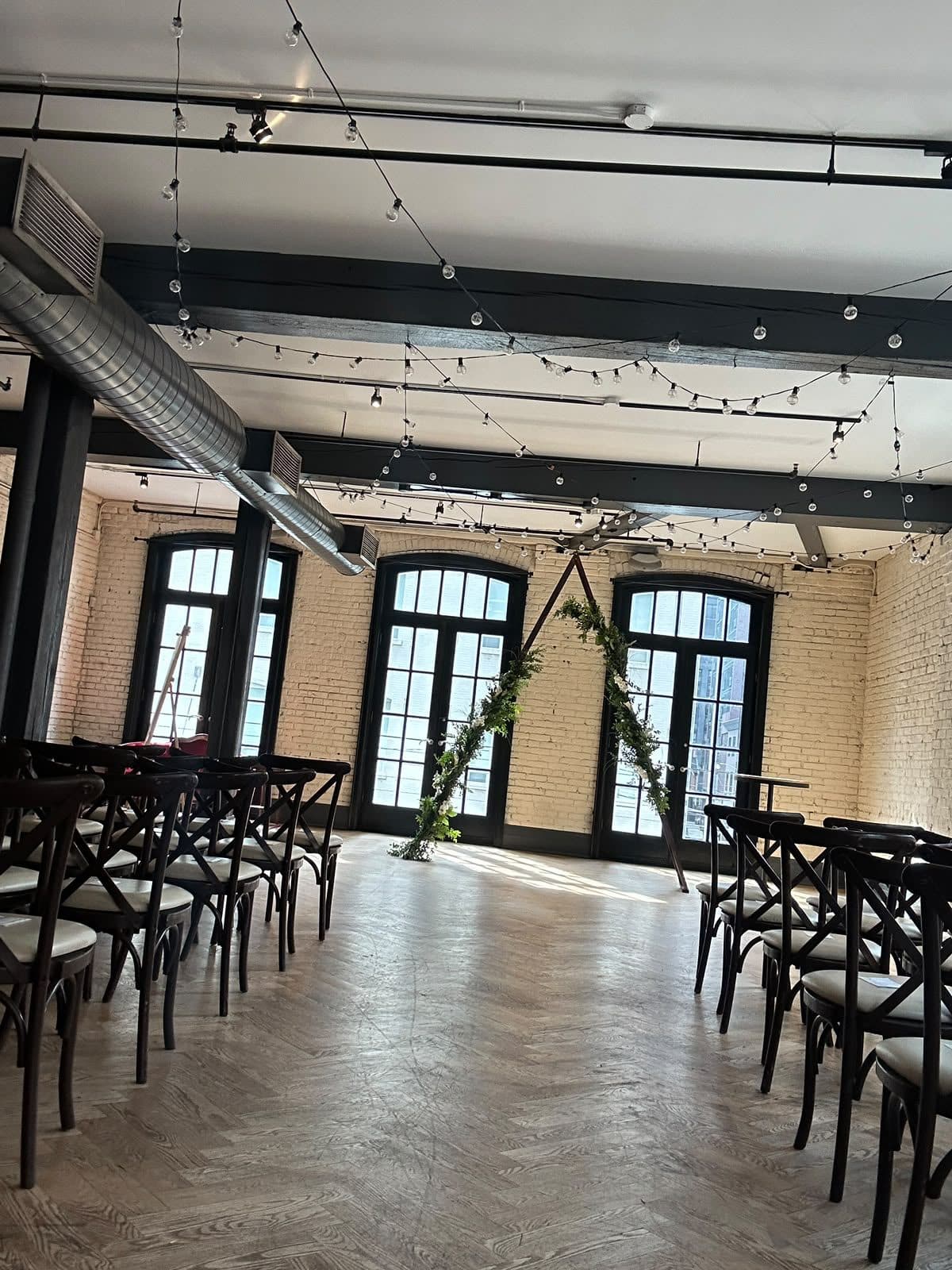 The same room set for the ceremony: rows of crossback chairs either side of an aisle leading to a greenery arch