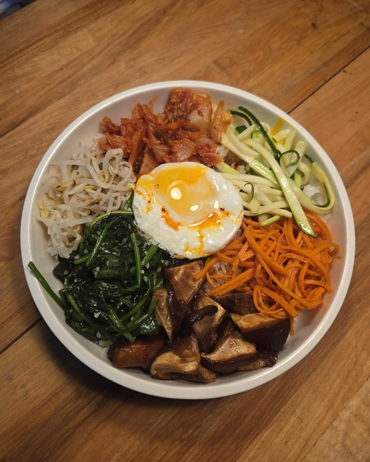 Bibimbap arranged in a bowl with rice, shiitake, kimchi, zucchini, seasoned spinach and a fried egg.