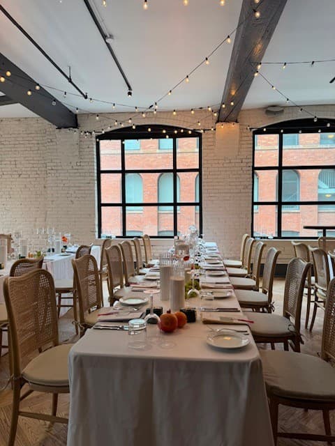 Second floor set for dinner: a long table dressed in white running the length of the room beneath tall arched windows, with festoon lights strung overhead and cane back chairs on either side.