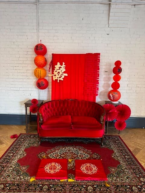 The tea ceremony backdrop photographed straight on: a red banner bearing the double happiness character, flanked by hanging paper lanterns and red paper fans, with two embroidered red cushions placed on a patterned rug in front of a red velvet settee.