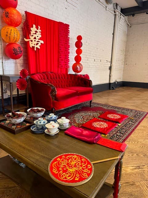The tea ceremony setting on the second floor: a red velvet settee against a red double happiness banner, paper lanterns in red and orange, floor cushions on a patterned rug and a tea service laid out on a low wooden table.