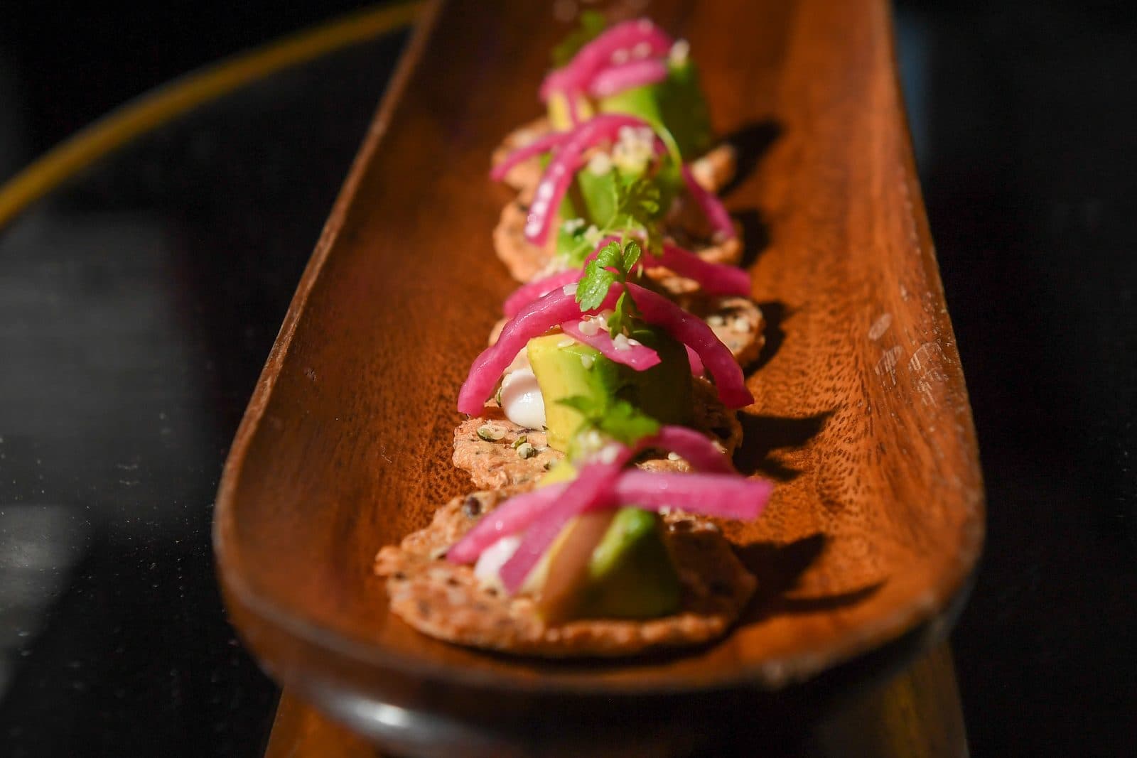A row of mini avocado bites on a long wooden board: wild rice crackers topped with avocado, pickled red onion and herbs.