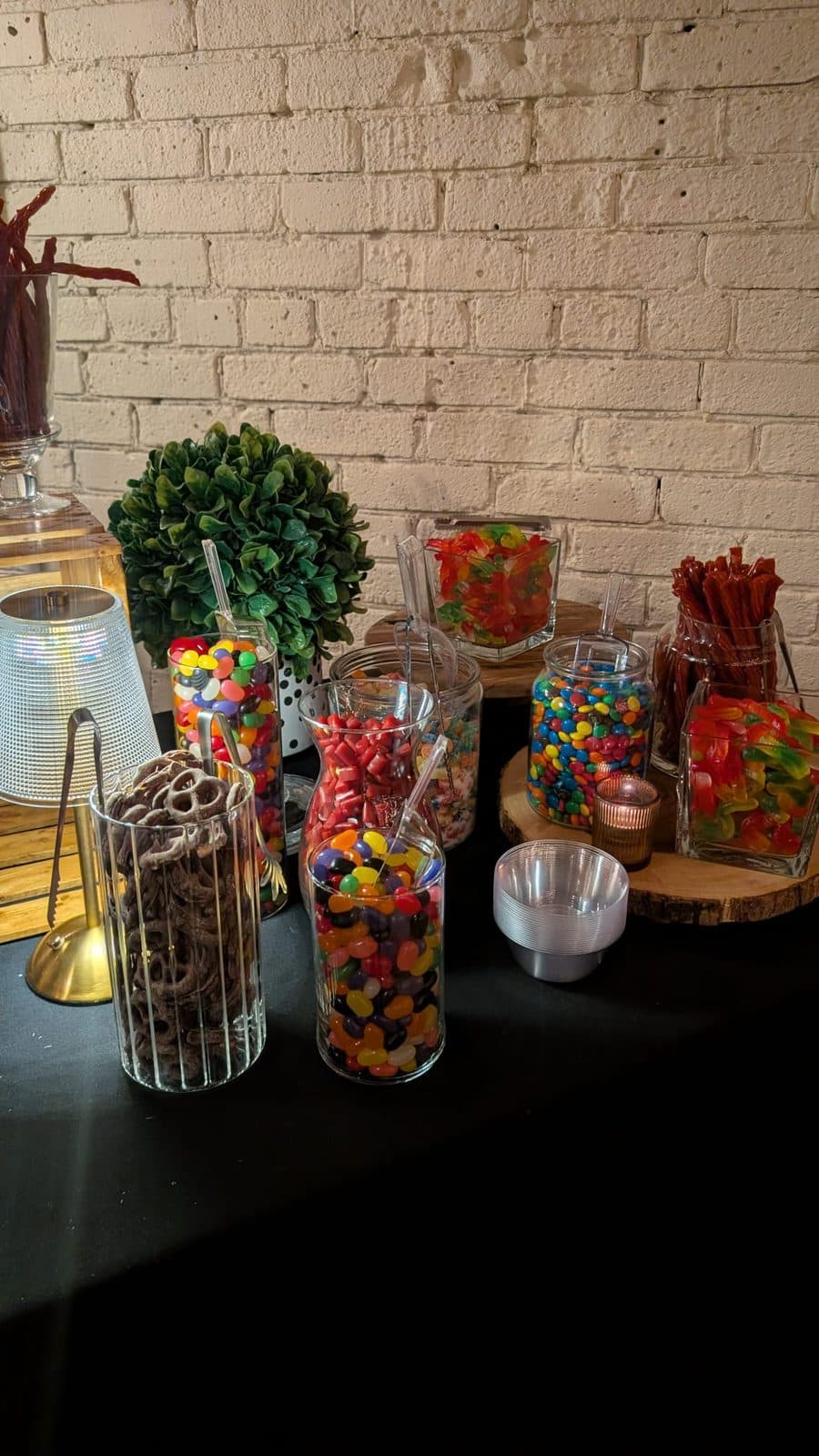 Tall glass jars of jelly beans, chocolate pretzels and gummies on wooden rounds against white brick, with a stack of small bowls beside them.