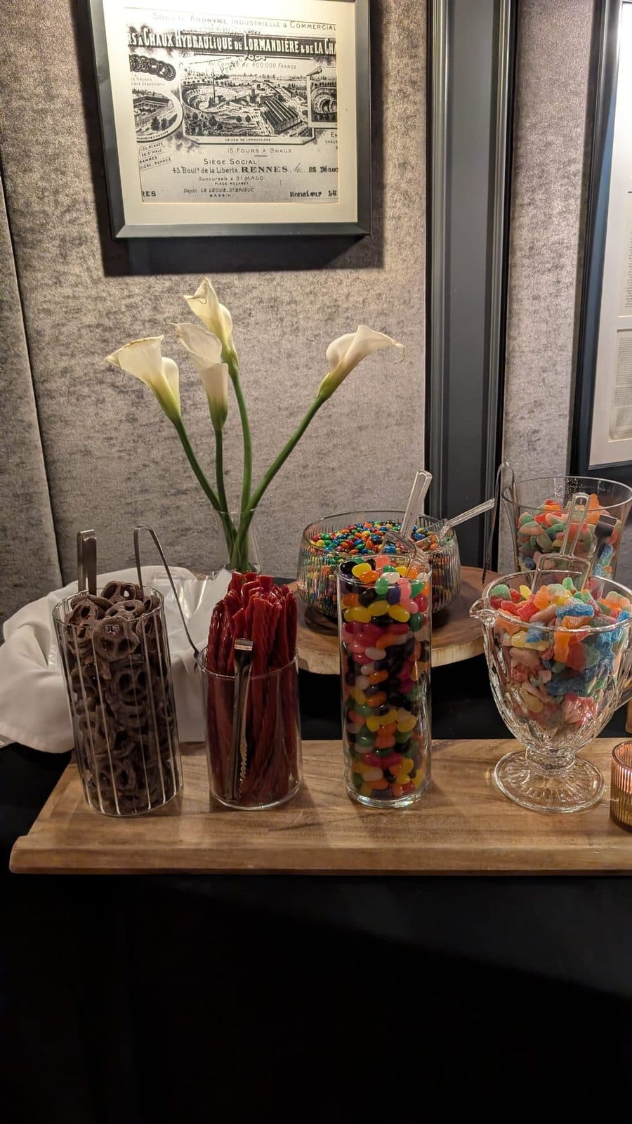 Glass jars of chocolate pretzels, red licorice, jelly beans and sour gummies on a wooden board, with white calla lilies in a vase behind them.