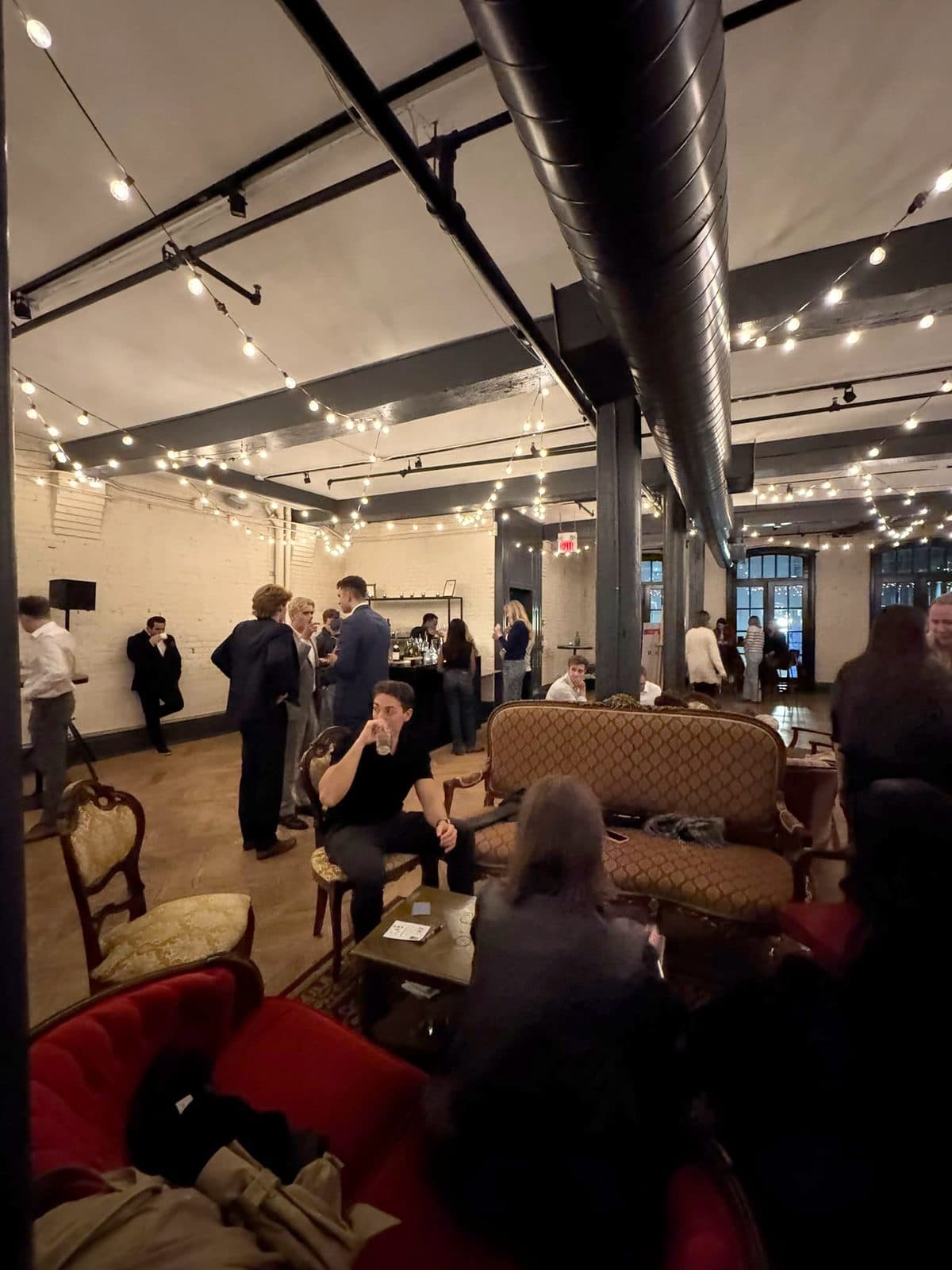 One of the lounge tables on the second floor: a tufted sofa, velvet armchairs and a coffee table, with guests standing and talking beneath the string lights.
