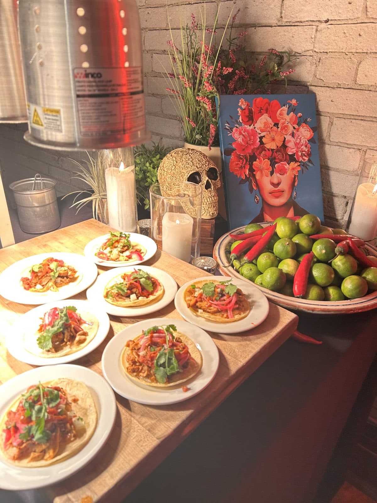 Plates of tacos topped with salsa and herbs laid out on a wooden counter, beside a bowl of limes and red chillies.