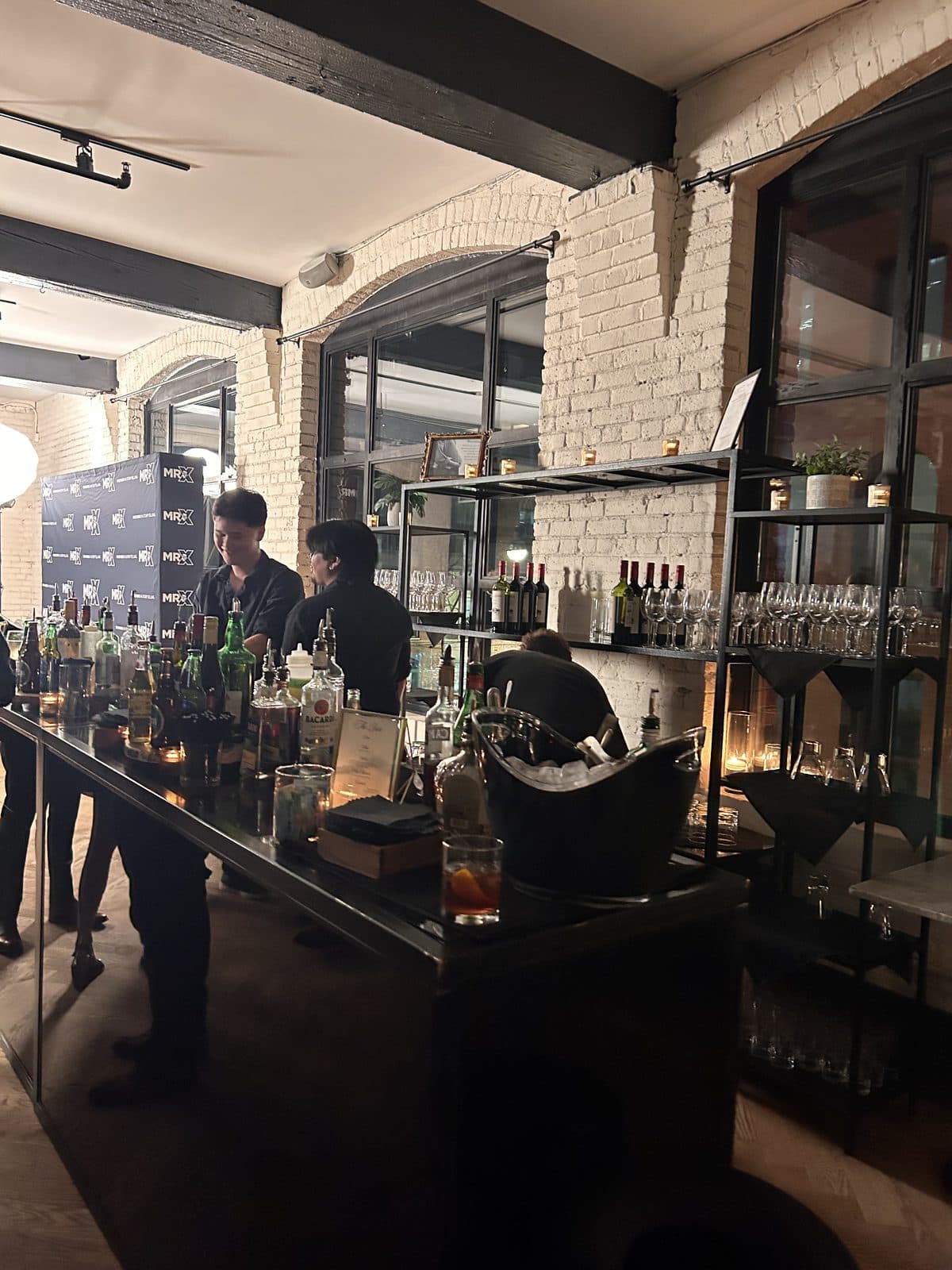 Bartenders working behind a long bar lit by candles, with bottles on open shelving and the arched windows behind.