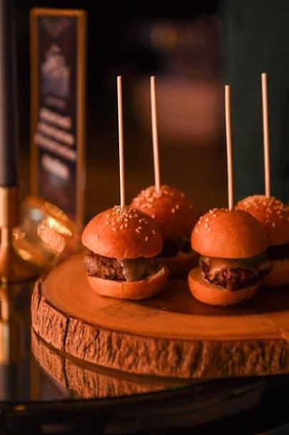 Small Storys burgers on sesame buns, skewered and set on a round wooden board under warm light.