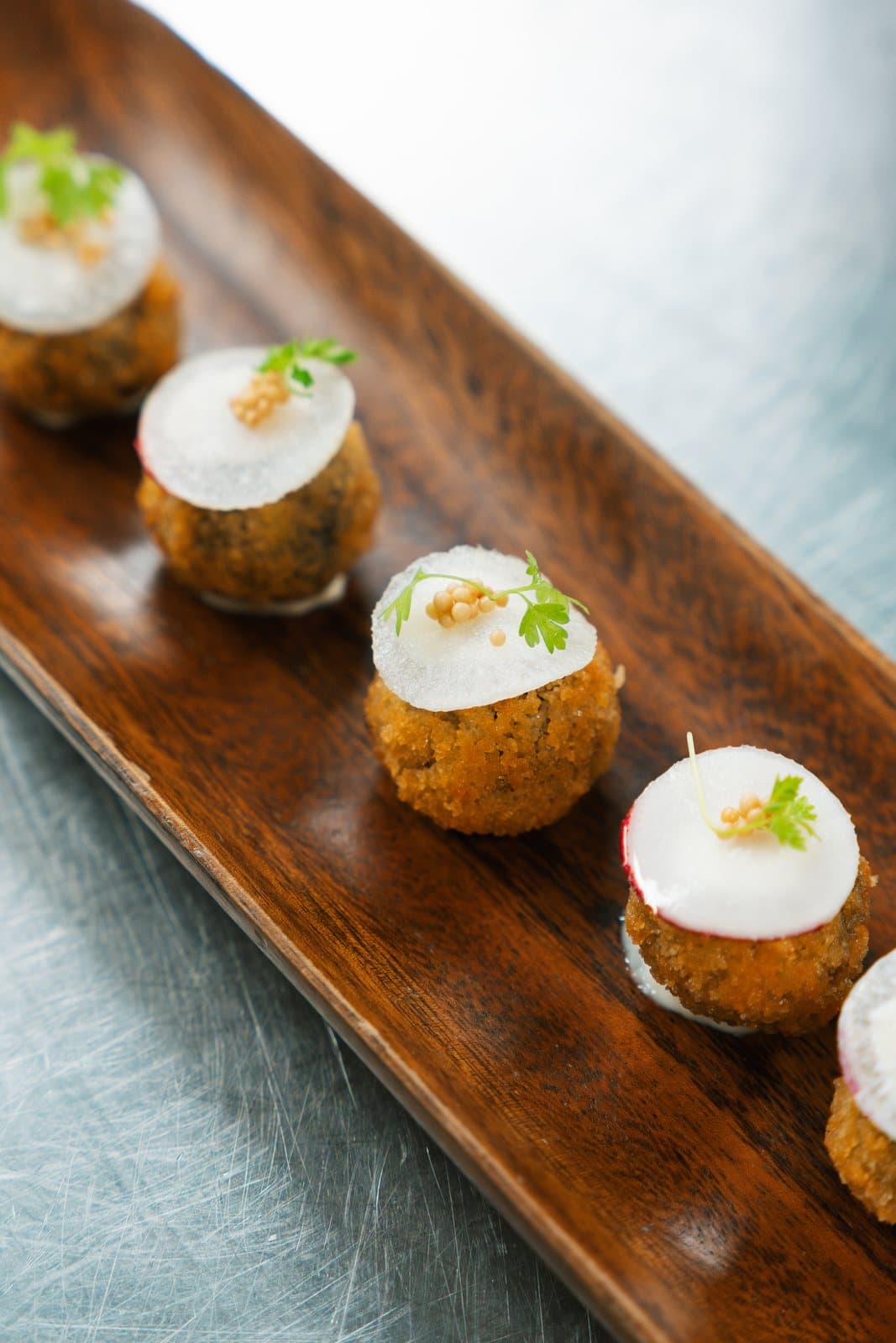 A line of round short rib croquettes on a wooden board, each topped with a disc of horseradish aioli and a sprig of herbs.