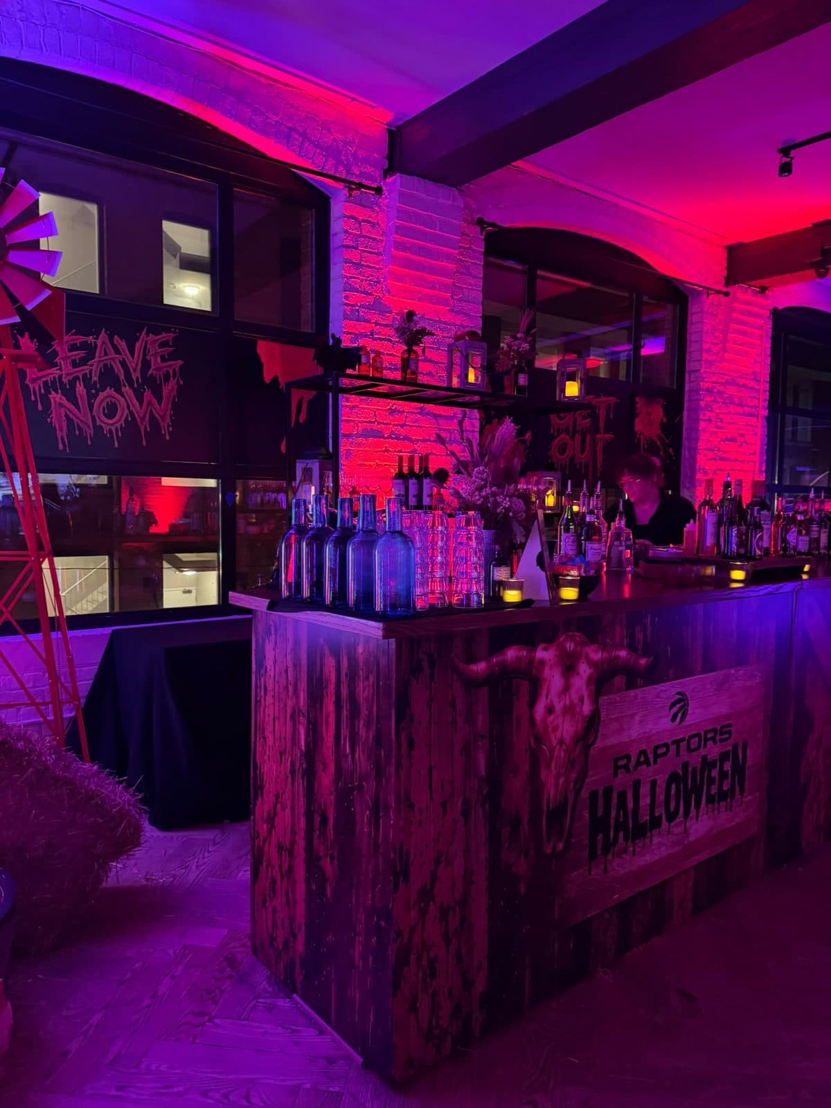 The bar wrapped in dark barn wood with a bull skull and a Raptors Halloween sign on its front, bottles lined up on top, and a Leave Now sign in the arched window behind.