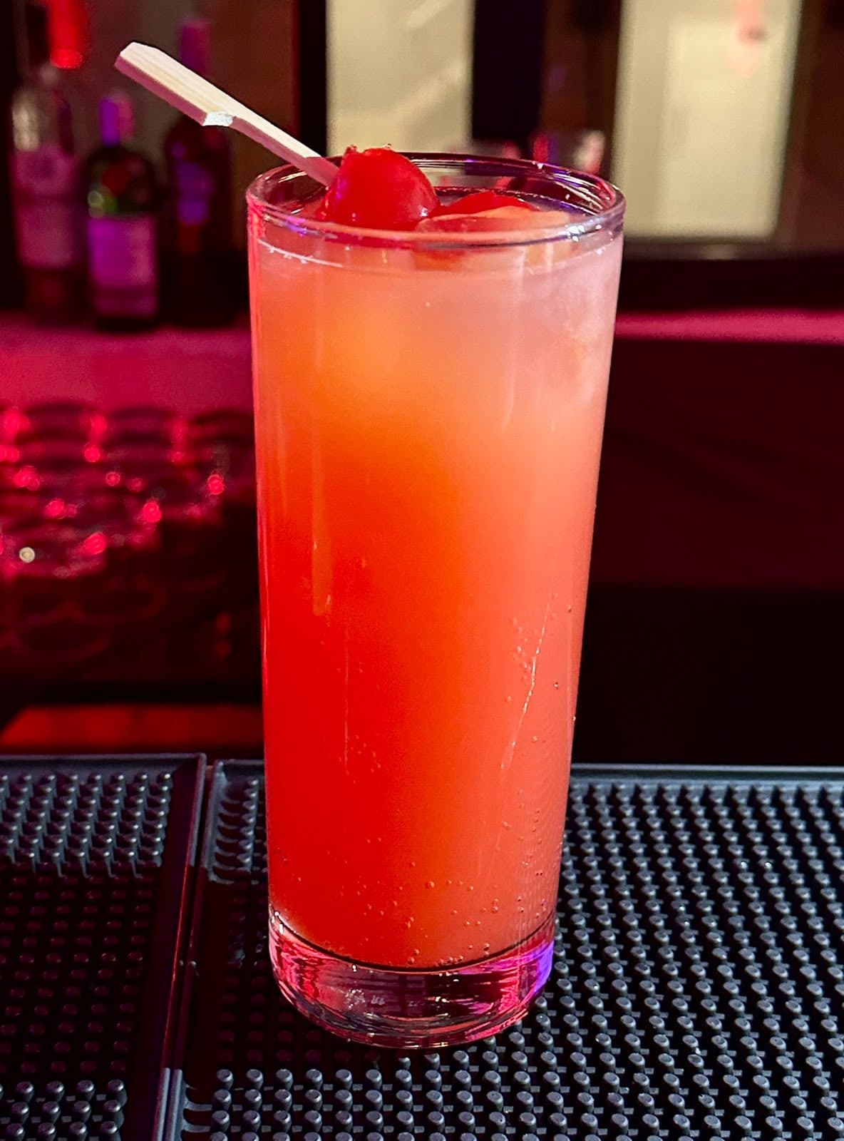 A tall red and orange cocktail with cherries and a pick, on a black bar mat.