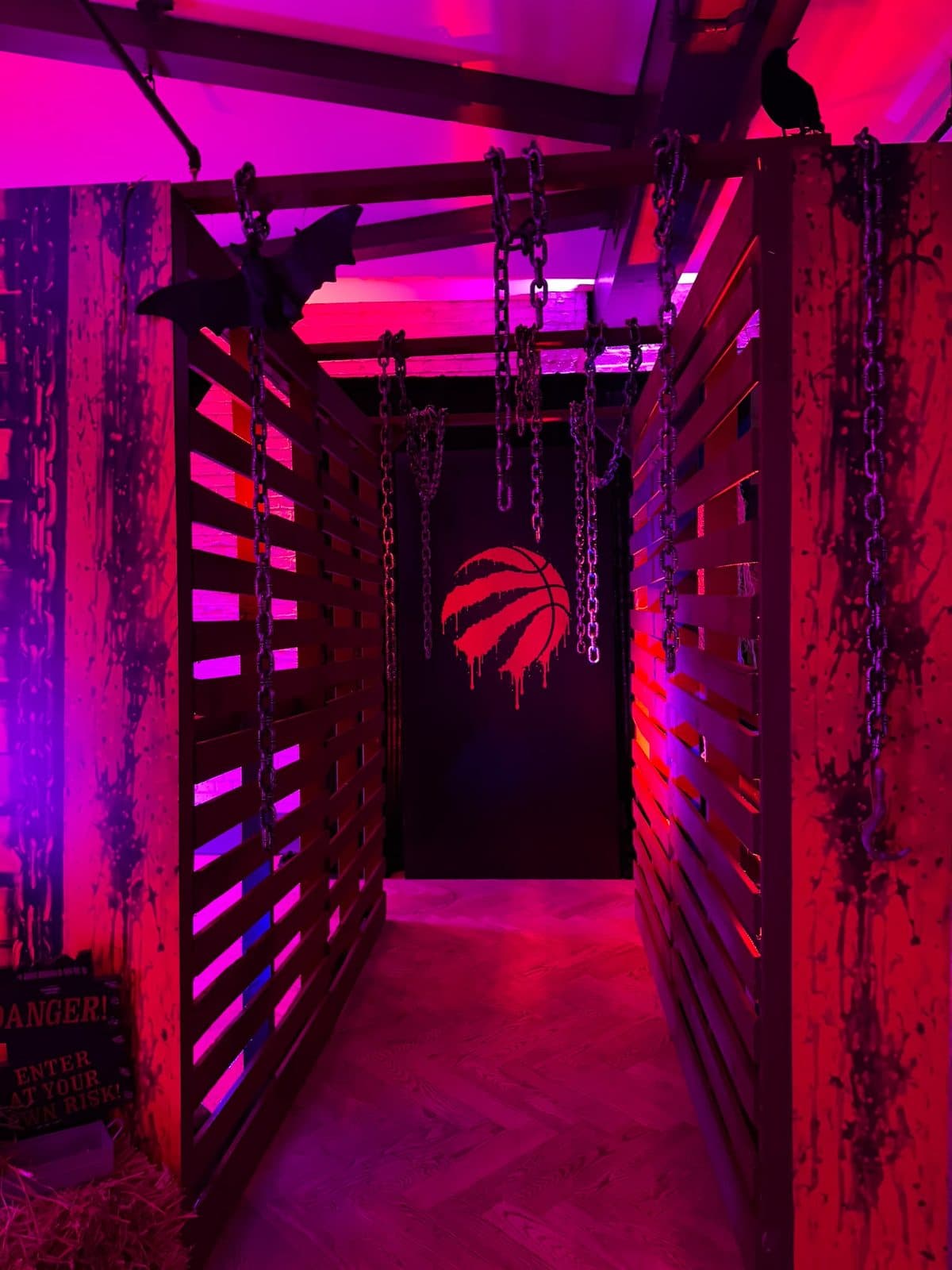 A corridor of slatted barn wood walls lit red and violet, chains hanging from the beams, a bat and a crow on top, a Danger sign in the hay at its mouth, and the Raptors claw at the far end.