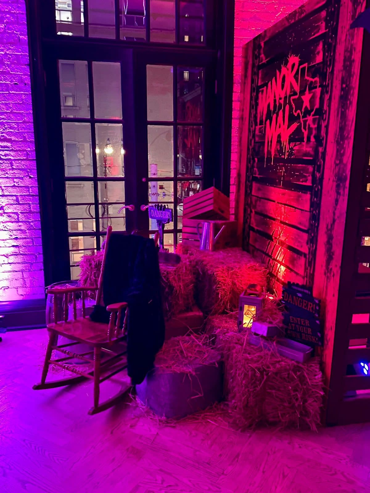 A rocking chair with a coat over it, hay bales, crates and a lantern beside a weathered wooden sign painted Manor Mal with an outline of Texas, in front of tall French doors.
