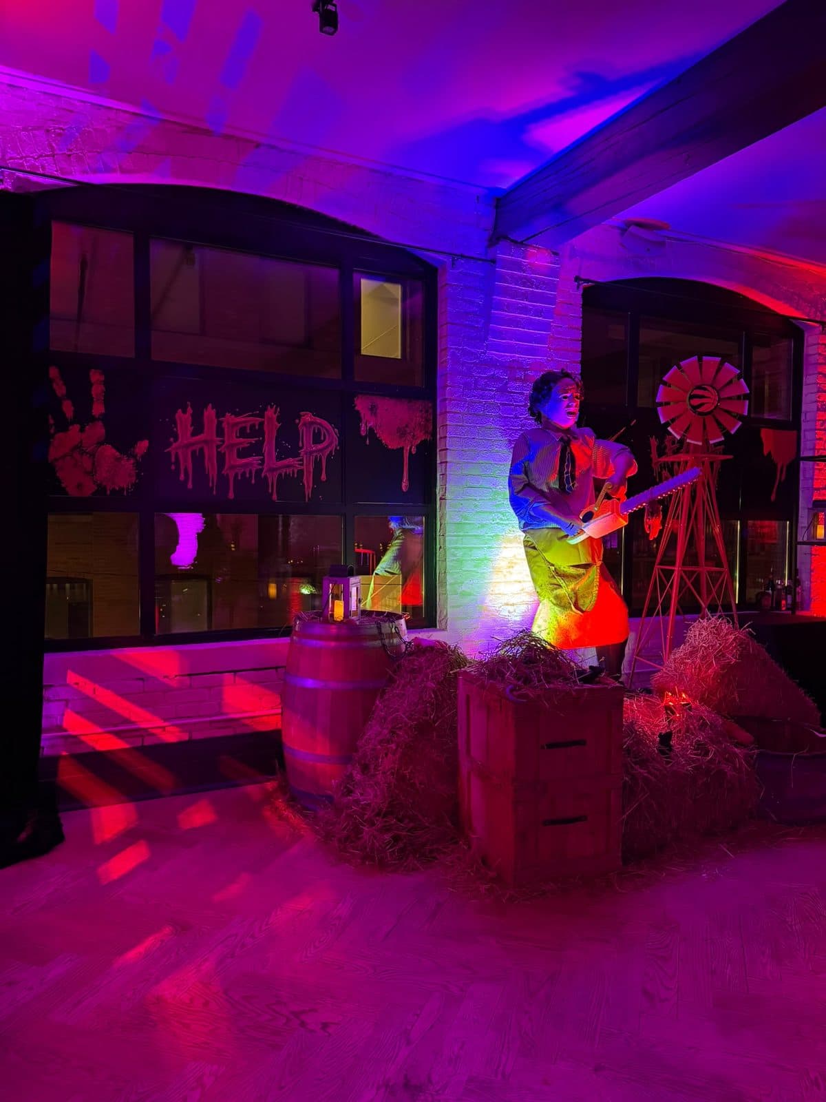 A life size figure holding a chainsaw stands on a wooden crate among hay bales and a barrel, a small windmill beside him, with Help and a bloody handprint painted on the arched windows behind.