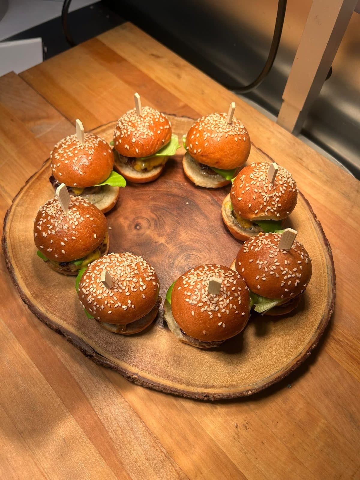 A ring of small sesame bun burgers on a round wooden board.