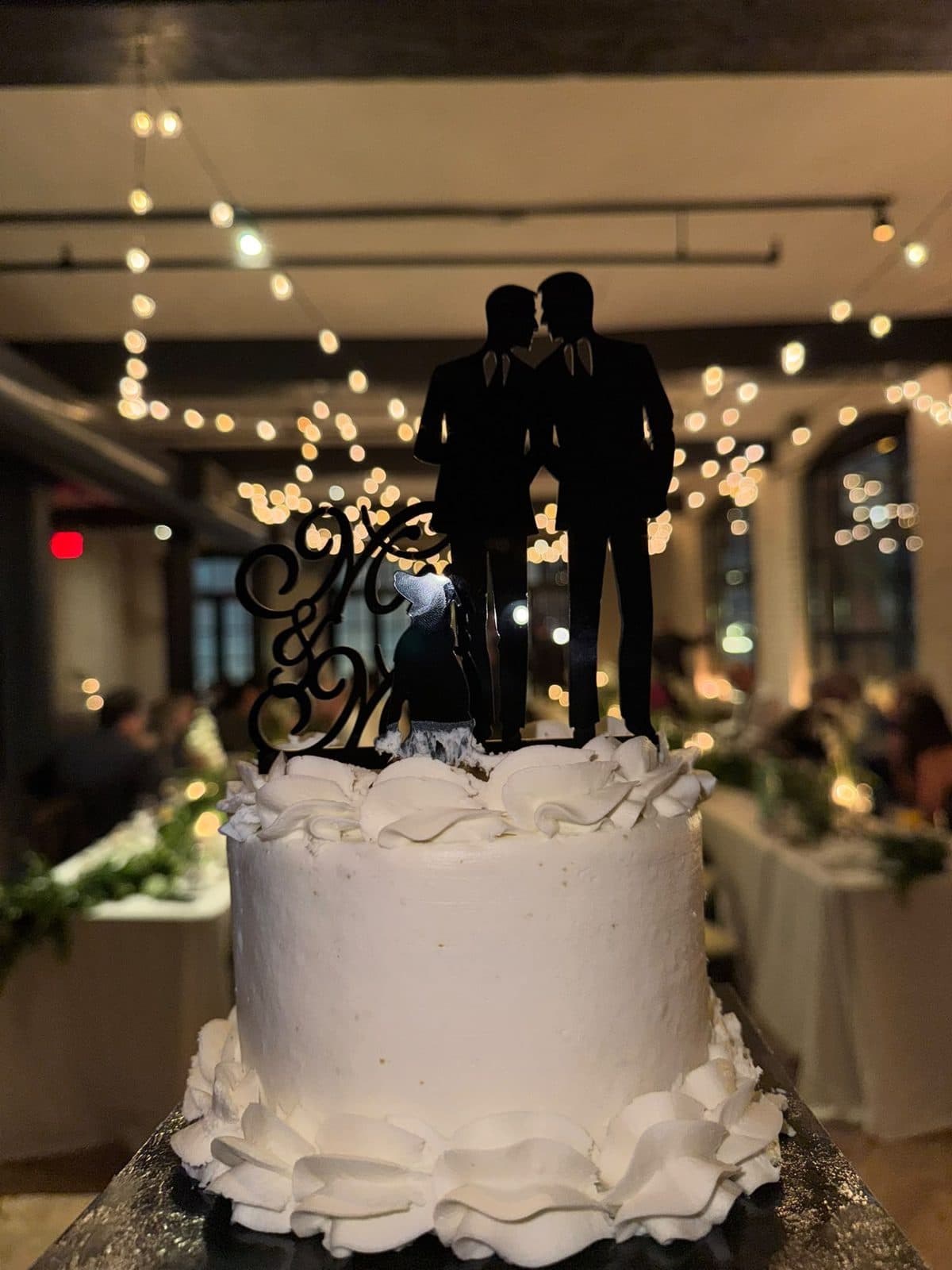 The small white cake with ruffled icing and a silhouette topper of two grooms, the dinner room glowing with string lights behind it.