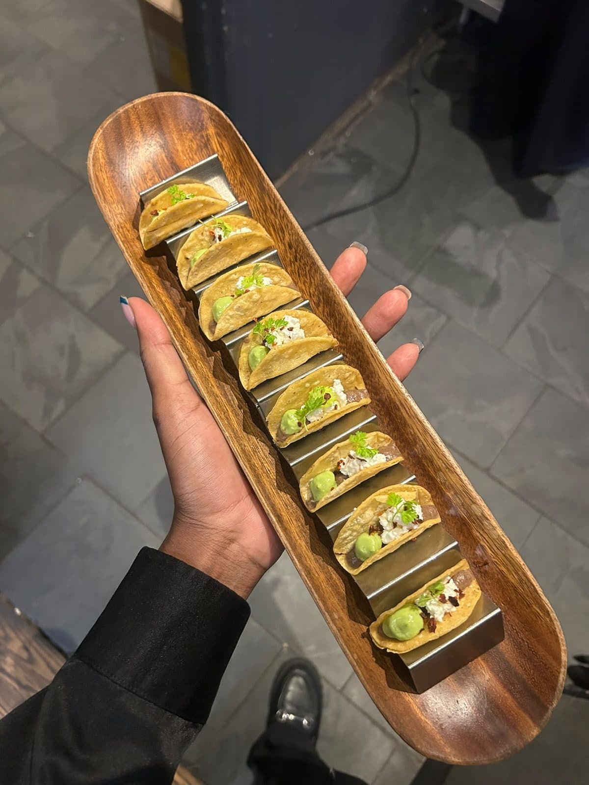 A row of small corn tacos held upright in a metal rack on a wooden tray.