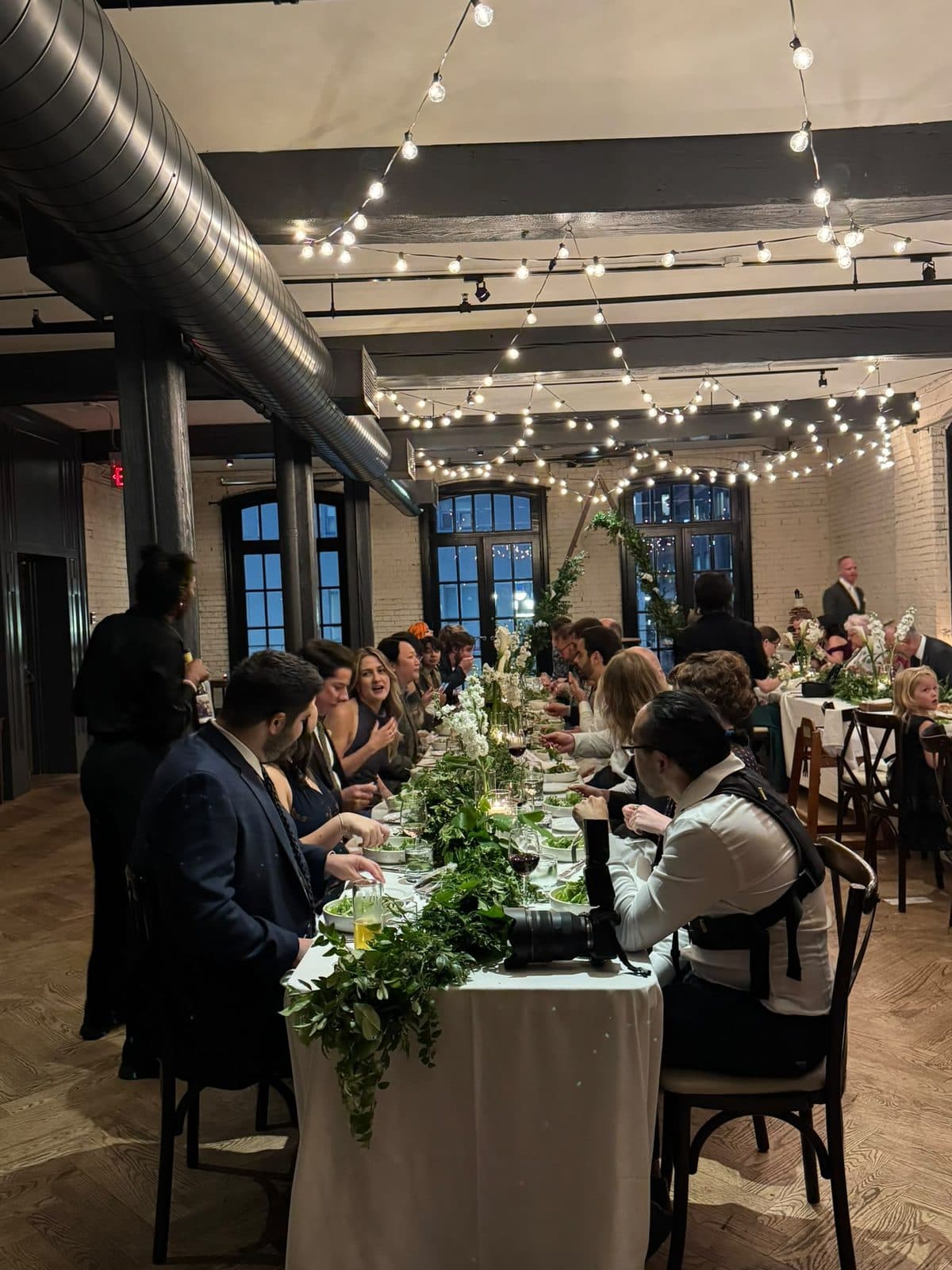 Guests at one of the two long dinner tables on the second floor, a garland of greenery running down the white tablecloth under strings of lights.