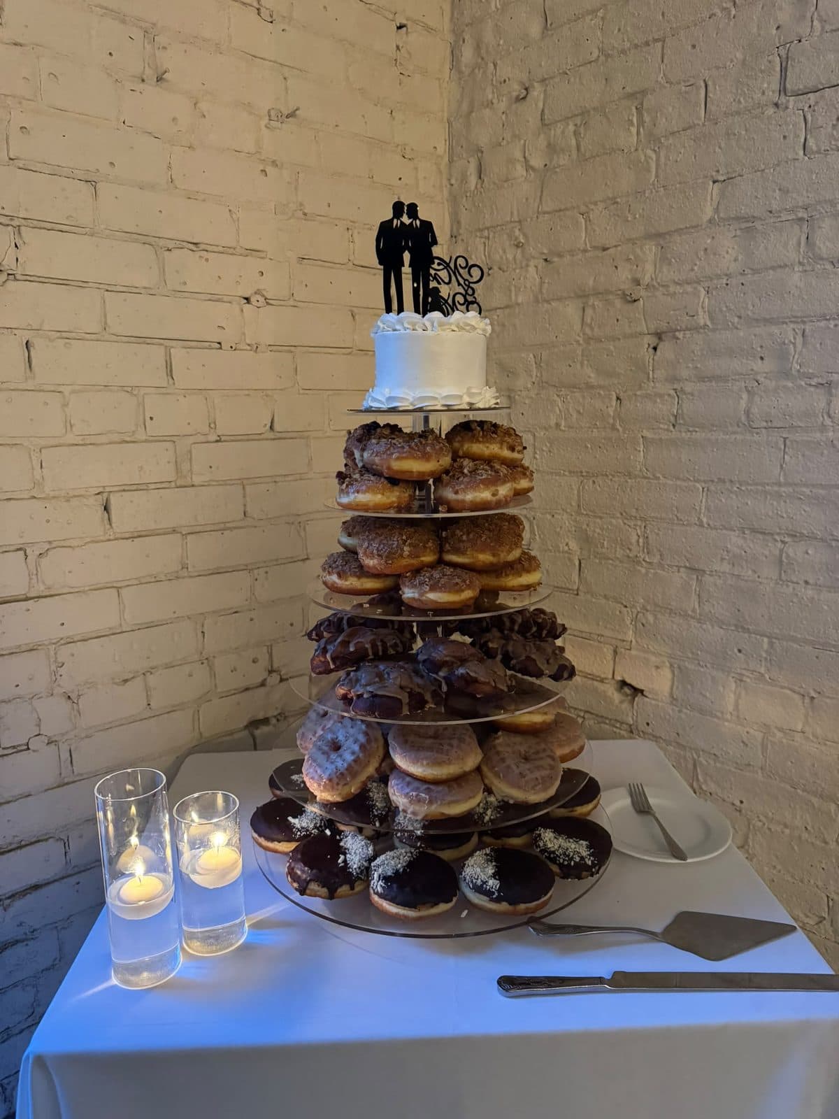 A tiered stand stacked with glazed and chocolate doughnuts, a small white cake with a silhouette topper of two grooms at the top, beside candles on a white table.