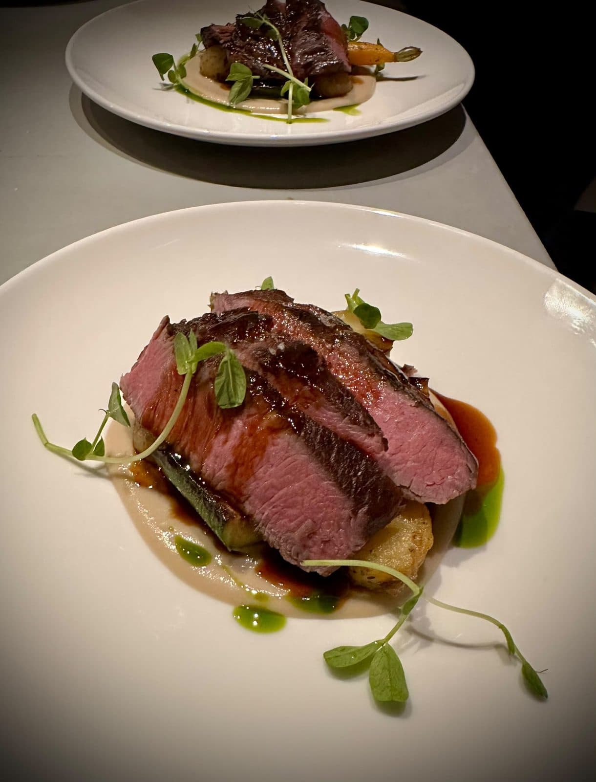 Sliced flat iron steak, pink in the middle, over potato and courgette with a dark jus and pea shoots.