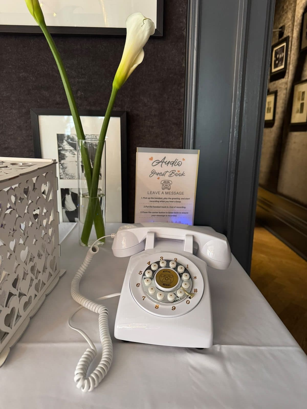 A white rotary telephone on a table with a small sign reading Audio Guest Book, leave a message, and a calla lily in a glass vase.