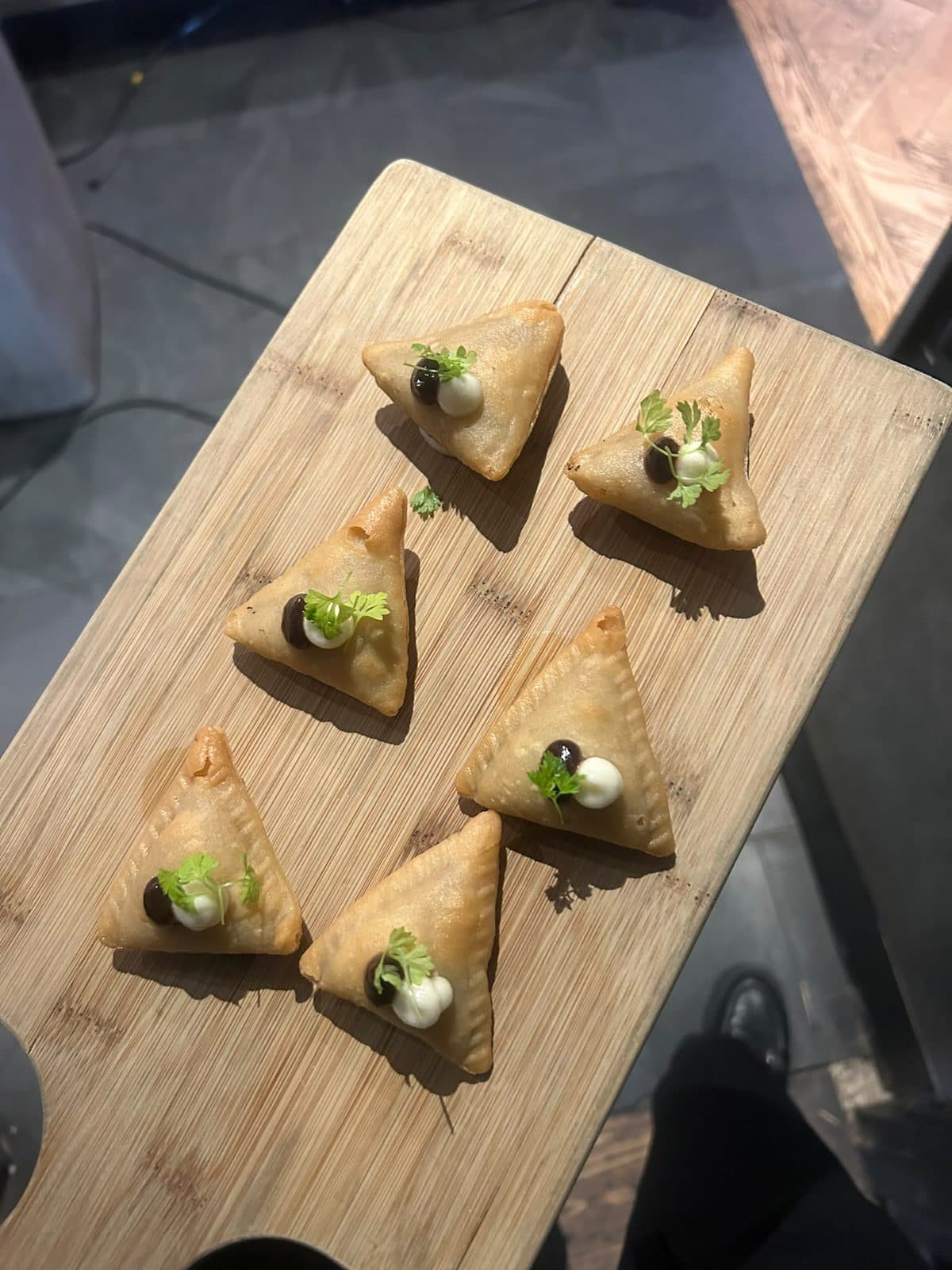 Small triangular samosas on a wooden board, each with a dot of sauce and a herb leaf.