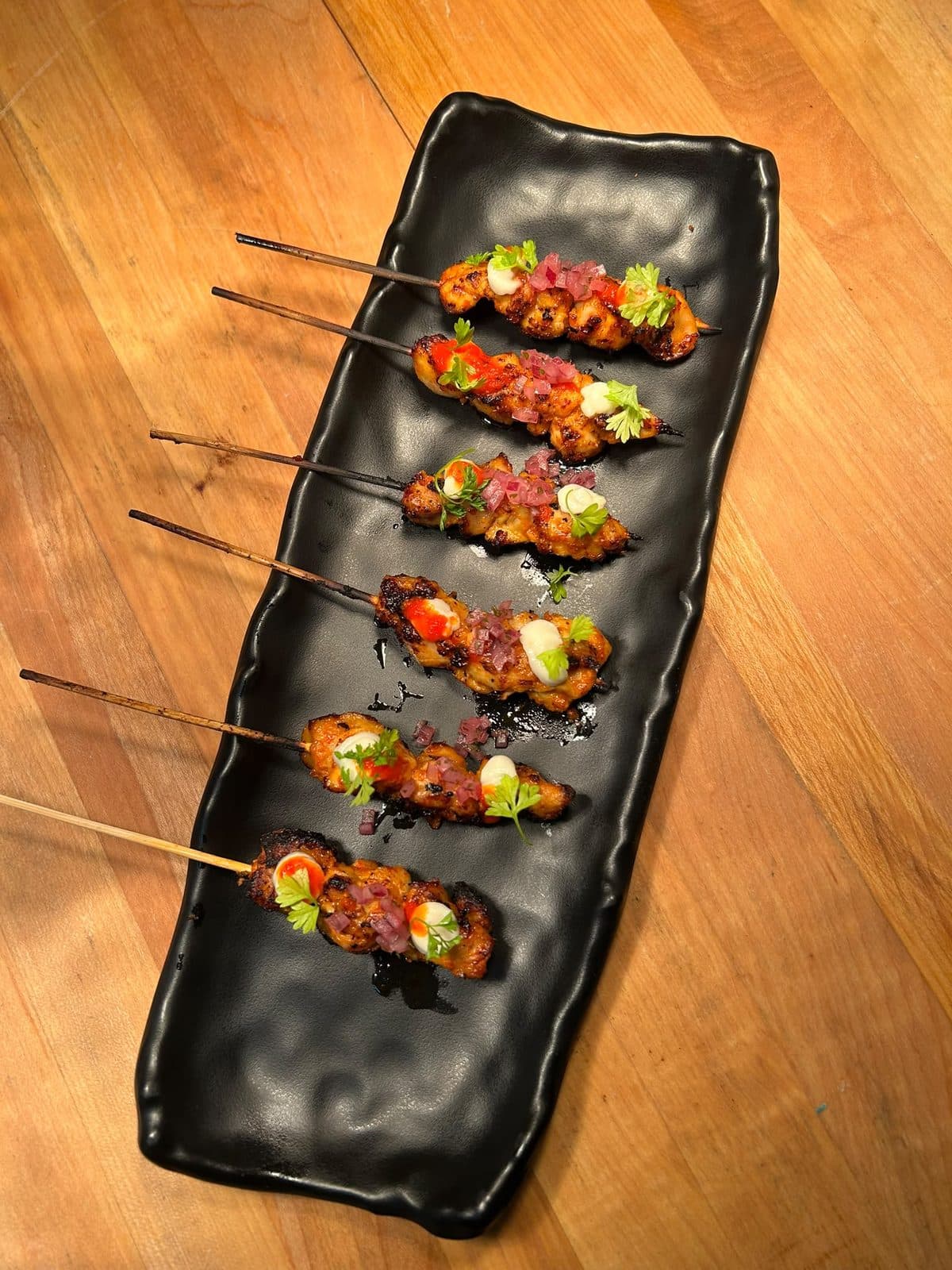Chicken satay skewers laid out on a long black platter.