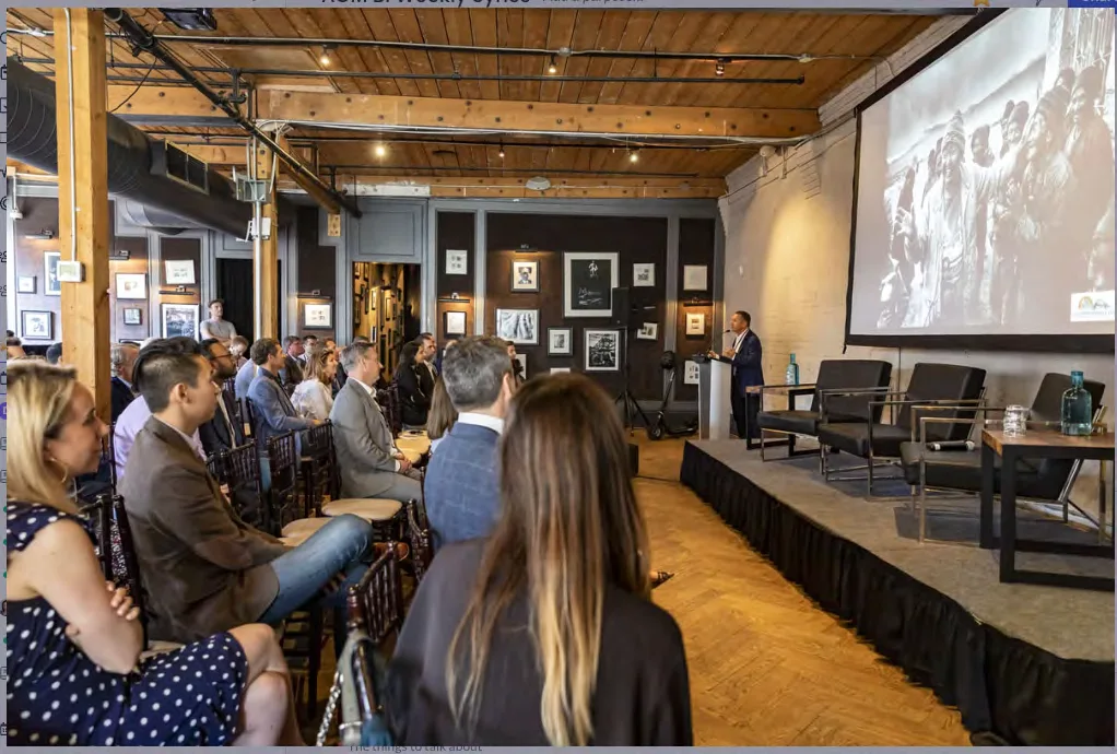 Professionals attending a corporate seminar in a modern event space with wood-beam ceilings, framed artwork, and a presentation stage with projection screen.