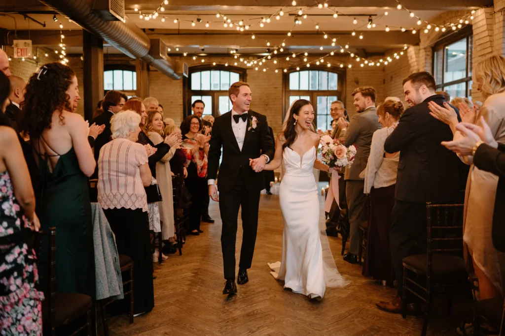 Just married couple walking down the aisle during an indoor wedding ceremony with guests clapping and fairy lights overhead