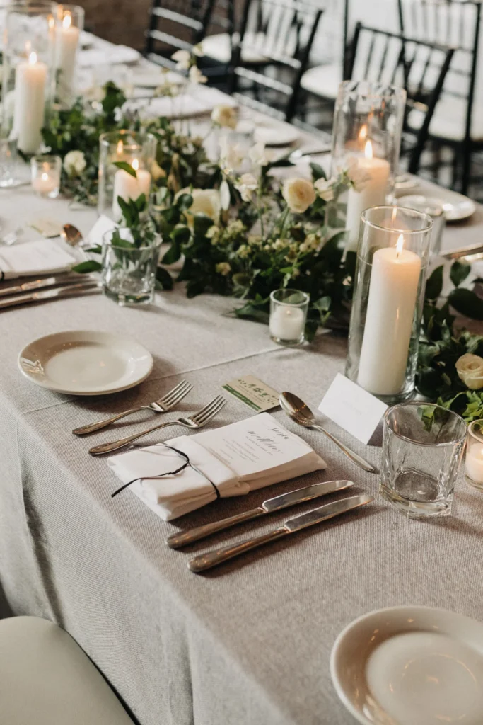 Wedding reception table setting with candles, floral greenery, custom menus, and a linen tablecloth.