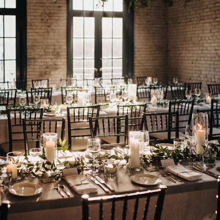 Beautifully decorated wedding reception tables with candles, greenery, and black chairs set in a warm, sunlit indoor venue.